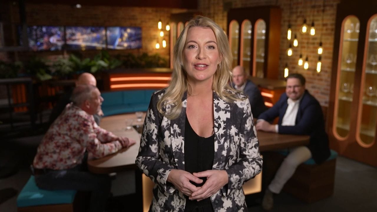 Presentatrice van De Bouwtafel in de studio, met gasten aan de tafel op de achtergrond
