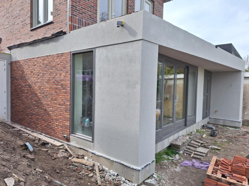 Hoek van de aanbouw in Amsterdam met grijze sierpleister en glazen puien, naast de bakstenen woning