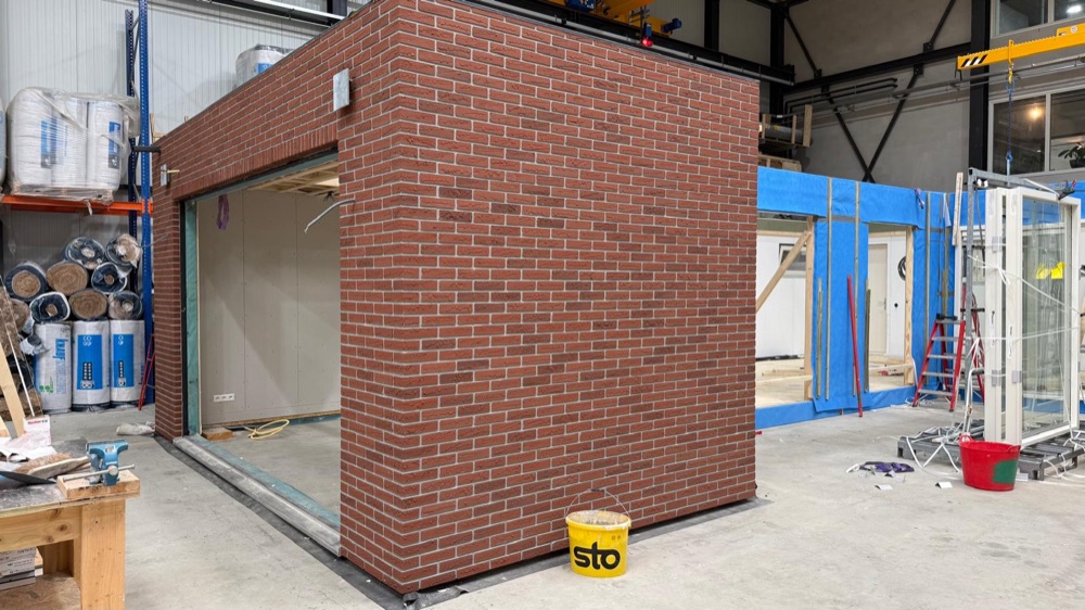 Prefab aanbouw met rode minerale steenstrips in de werkhal, met een emmer Sto ernaast