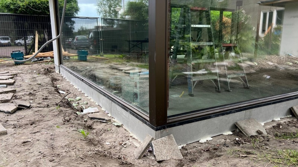 Glazen hoekpartij met donker kozijn op de plint, met grondwerk ervoor, bij een woonhuis in Amsterdam-Noord tijdens de uitvoering